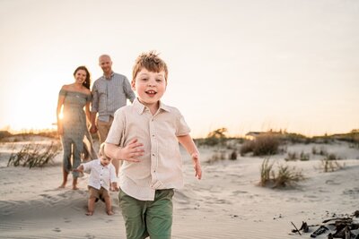 Little boy running towards the photographer with a playful smile, the rest of the family behind him are smiling at the camera