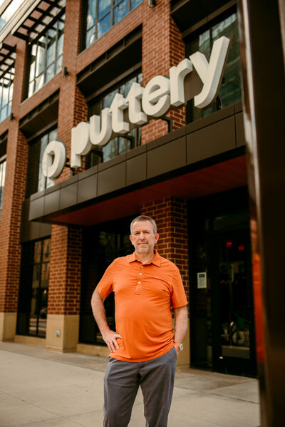 Outdoor professional dating profile photo of Kerry in Minneapolis, standing in front of a restaurant in a bright orange shirt, confident and natural.