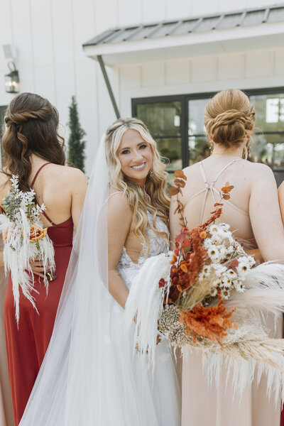 Bride and bridesmaids lined up in front of chapel, bridesmaids face the other way as bride looks back towards camera and smiles with bouquet in hand