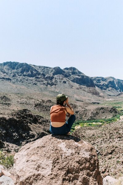 Person sitting on a large rock overlooking the rugged desert landscape of Big Bend, capturing the view with a camera — Big Bend elopement photographer capturing scenic moments in nature.