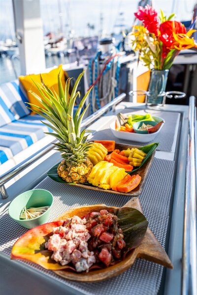 Close-up of tropical food on a Hawaii sailboat charter, including fresh pineapple, sliced fruit, and poke served on a tray beside blue striped seating.