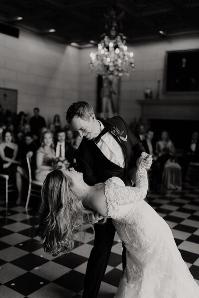 Groom dips the bride during their first dance during this classy wedding at the Marland Mansion in Oklahoma as their family and friends surround them with champagne and happy tears