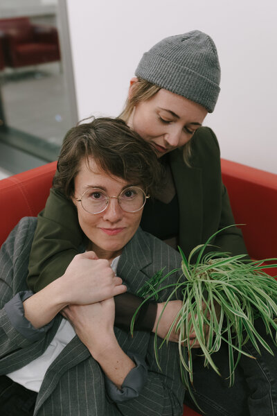 Two women embrace on a couch while holding a spider plant bouquet after their wedding at City Hall - Courthouse elopement in Portland, Oregon. 