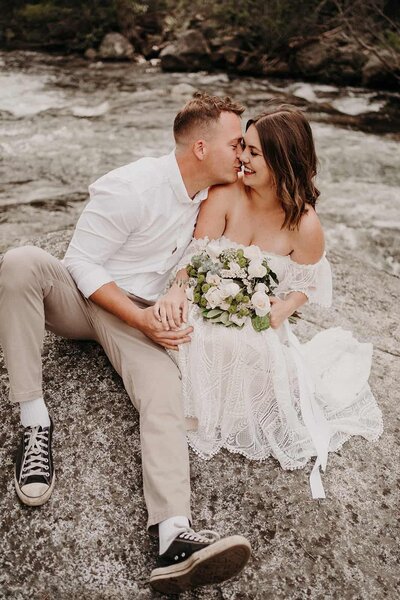 a bride stroking a groom's face while standing on a cliff side
