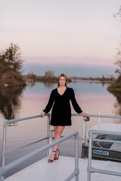 A senior girl poses for the camera in a little black dress on the dock of a lake.