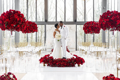 Bride and groom in white standing on a circular bed of red roses, surrounded by towering rose arrangements and candlelit aisle inside a modern glass chapel, designed by Wells Design Co