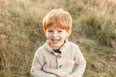 red-haired boy giggling at the camera. He is wearing a cream sweater and out in nature surrounded by tall golden grass.