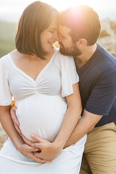 Pregnat womand and her husband, close to each other, forehead to forehead, smiling and holding the belly. Sunset light around them. 