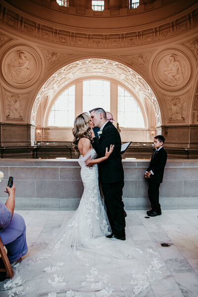 Bride and groom sharing their first kiss during an intimate elopement ceremony at San Francisco City Hall, with guests and children looking on, captured by San Francisco elopement photographer Kristin Smith.