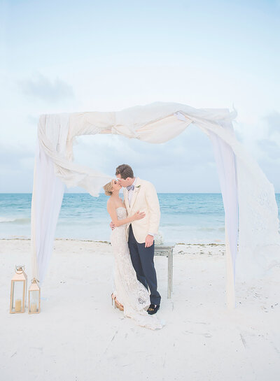 Bride and groom kissing under white curtain arch on beach