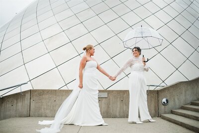 Two brides in elegant gowns holding hands beneath a white umbrella at the Museum of Glass in Tacoma, captured by Seattle wedding photographer Candace Connor