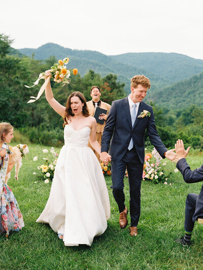The bride cheers with her bouquet raised and the groom high fives a guest as they exit the ceremony, during their summer wedding in North Carolina, by destination photographer My Sun and Stars Co.