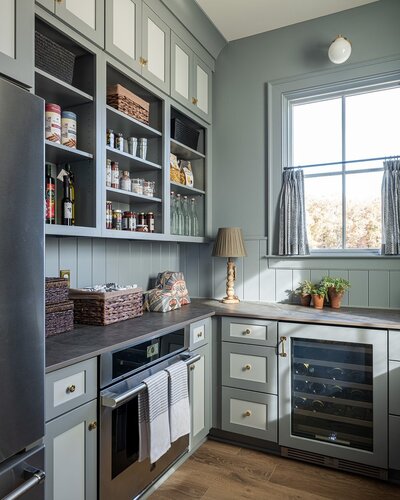 English Nautical Pantry and Modern Minimal Walnut Bar Los Angeles Kitchen. This custom-designed bar and scullery space merges sophistication with practicality, offering a seamless transition between high-end aesthetics and daily functionality. The color palette incorporates elegant gray-blues and walnut wood finishes, while the built-in storage maximizes space efficiency. The design features integrated appliances, sleek cabinetry, and custom shelving to create an organized yet stylish area for entertaining and home organization.