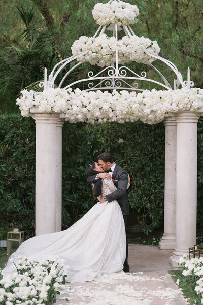 A newlywed couple shares a passionate first kiss under an elegant floral-adorned gazebo at The Beverly Hills Hotel. The bride’s stunning gown flows behind her as she leans into the embrace of her groom, surrounded by lush greenery and white roses. Captured by an LA wedding photographer.