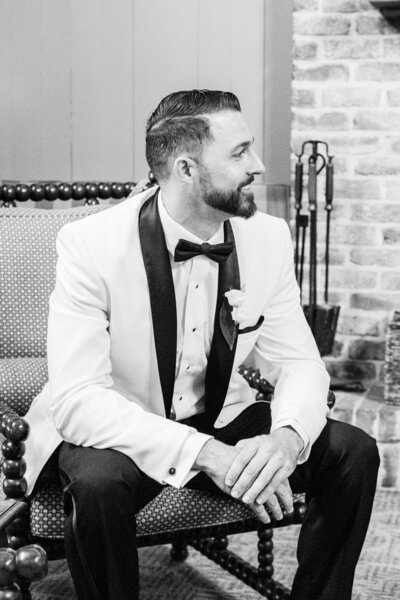 A groom in a crisp white tuxedo with black satin lapels and a classic bow tie sits in a refined lounge at Montage Palmetto Bluff, exuding timeless elegance and anticipation on his wedding day.