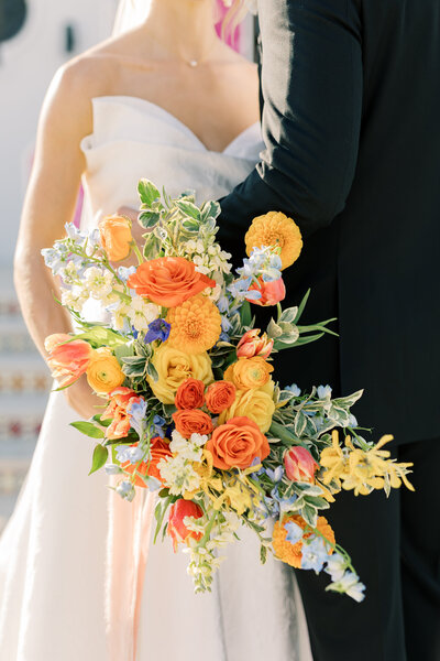 A bright, colorful bridal bouquet with orange tulips, yellow ranunculus, orange dahlia’s yellow orchids, blue delphinium, and yellow roses