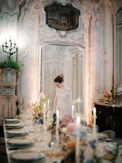 A bride in a Galia Lahav lace dress stands in a beautifully decorated room in Villa Sola Cabiati with ornate walls and a large mirror. There is a table set for a formal dinner with lit candles, floral arrangements, and fine tableware in the foreground.