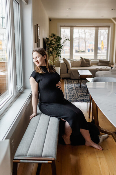 Pregnant woman dressed in fitted black dress sitting on a bench and casually looking to her sid out the window.