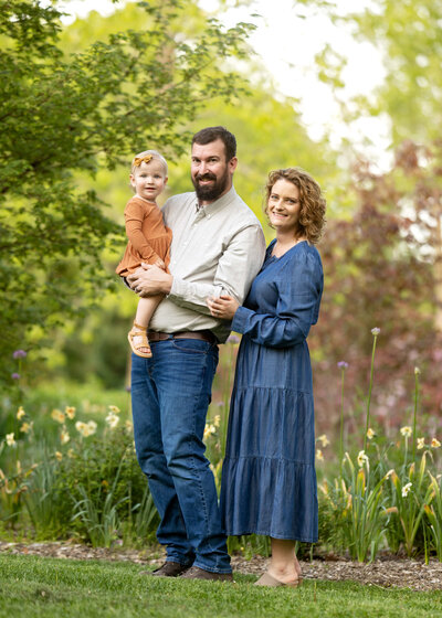 A family of a mom, dad and toddler girl are standing in the flowers, smiling