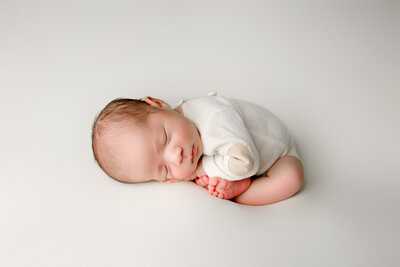 Newborn baby boy in womb pose wearig cream romper lying on white background with fresno newborn photographer