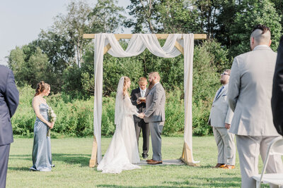 bride and groom holding hands under ceremony arch with bridal party and guests during wedding photo session in upstate ny 