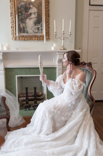 Bride in her wedding gown sitting and looking in a mirror at herself after services with Indianapolis makeup artists and hairstylists, Something Blue Stylists