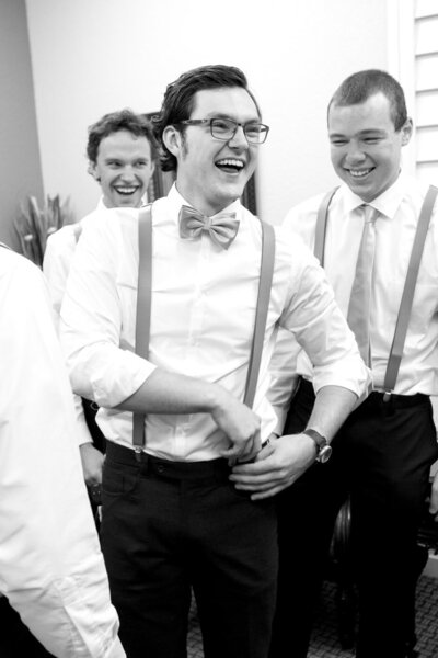 A man wearing suspenders and a bow tie laughs with two of his groomsmen.