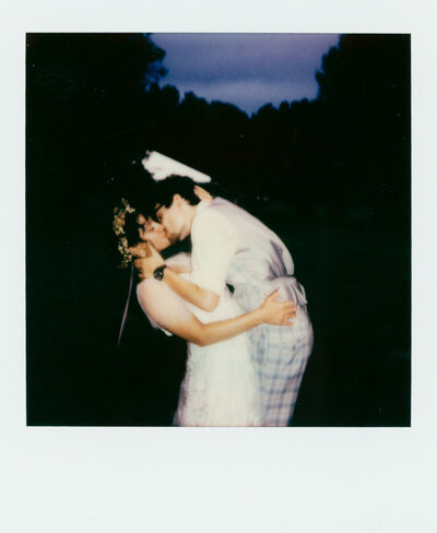 Polaroid photo of a newlywed couple kissing at dusk in a field, wearing playful wedding attire — a dreamy image captured by a film wedding photographer.