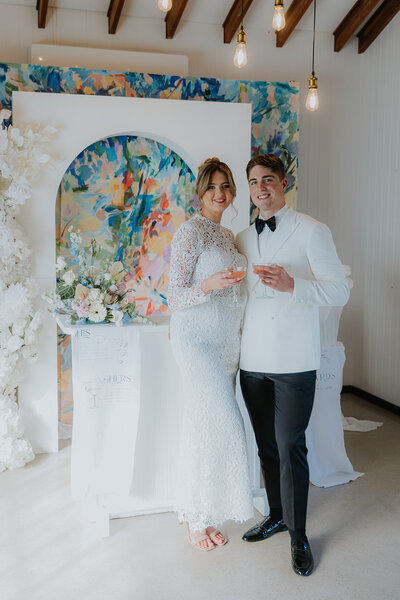 A man and woman hold cocktails whilst dressed in a white tuxedo and lace wedding dress. They are celebrating their Elopement on the Central Coast at Our Secret Garden with an Art Gallery theme. There is a colourful mural behind them and a mobile bar with flowers on top.