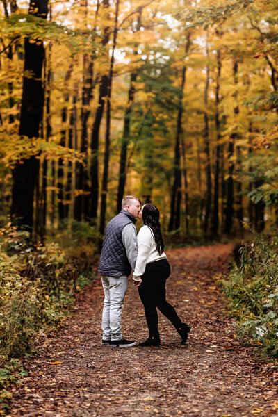 Holding hands, a couple shares a kiss along a path lined with beautiful fall foliage