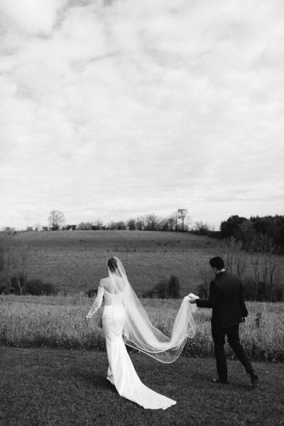 A couple in wedding attire walks across a grassy field with rolling hills under a cloudy sky, captured by Ontario destination wedding photographer, Jennifer van Son at Farmhill Weddings in Peterborough.