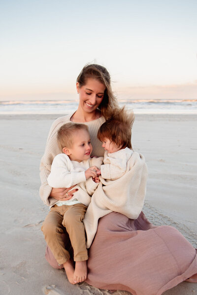 Smiling mother sitting on the beach with her two children wrapped in neutral knits, captured in a candid family photo session on Florida’s coast.