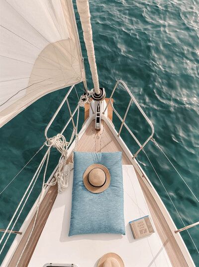 Peaceful sailing scene with a fedora and book on a sailboat bow, floating in turquoise water. The essence of slow, intentional travel. Tailored Oasis Travel Co.