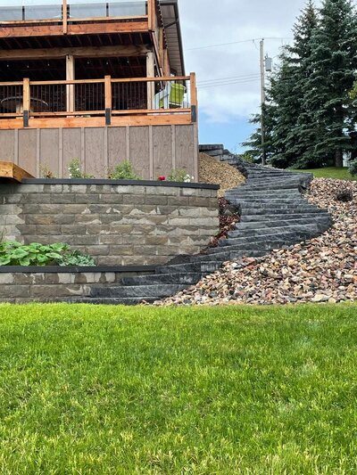 Sloped backyard with one retaining wall that is 6 feet, concrete steps to access the backyard from the deck, limestone garden beds with perennials ideal for Airdrie.