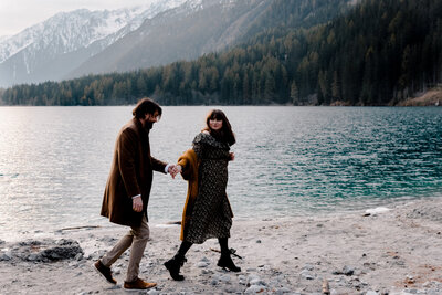 Frisch verlobtes Paar läuft entlang einem weißen Strand mit Bergkulisse im Hintergrund.