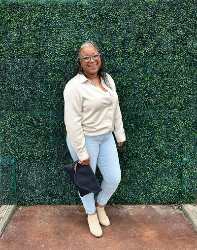 Letitia Hopkins standing confidently in front of a plant-filled greenery background, holding a stylish purse, embodying self-love, confidence, and Main Character Energy as a life, love, and relationship coach empowering women to create aligned, fulfilling lives and relationships.