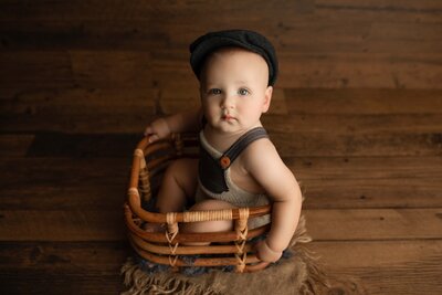 First birthday photoshoot in London, Ontario - Baby boy sitting in a basket wearing knit overalls and a paperboy cap looking at the camera. Baby is against a wood backdrop.