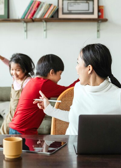 Busy mom working at laptop and appearing to try to calm hyper kids down so she can work.