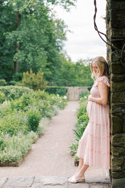 A pregnant woman wearing a pink floral dress leans against a stone wall in a garden during her NJ maternity photography session.