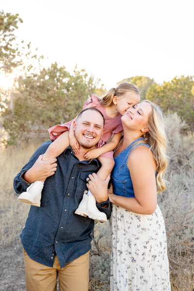 Little girl kisses moms cheek as she is sitting on dads shoulders.
