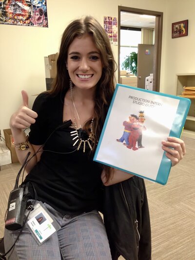 Madison proudly holding her “Production Intern” folder at Sesame Workshop, giving a thumbs up on her first day.