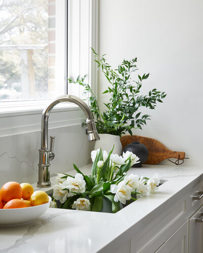 A kitchen sink with a polished nickel faucet surrounded by potted plants and fresh fruit, embedded in a marble countertop below a window