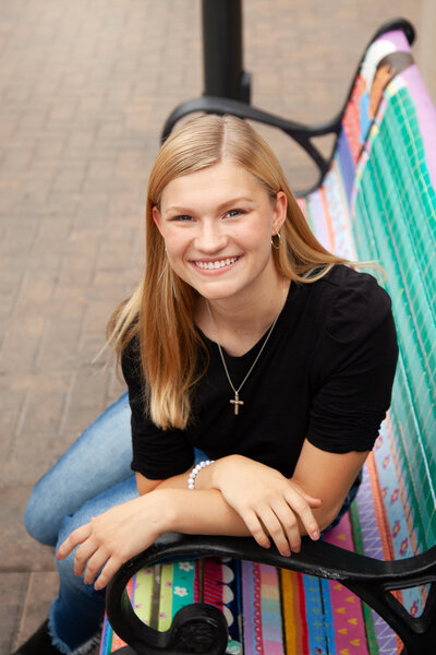 Senior girl wearing a cross necklace, sitting on a colorful bench, smiling at camera.