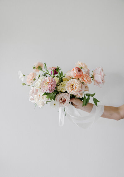 A soft romantic bridal bouquet in pastel tones featuring blush and peach garden roses, buttercream ranunculus, pale pink zinnias, lisianthus, and delicate greenery, tied with flowing white ribbon and photographed against a clean light-gray background for a modern fine-art wedding aesthetic.