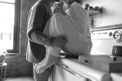 A woman is sitting on a stove with a man standing, embracing her.