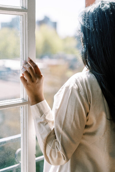 A person with long dark hair, wearing a light-colored shirt, stands by an open window and looks outside, resting one hand on the frame. Sunlight filters in—evoking the calm sought in anxiety and depression counseling Southern California.