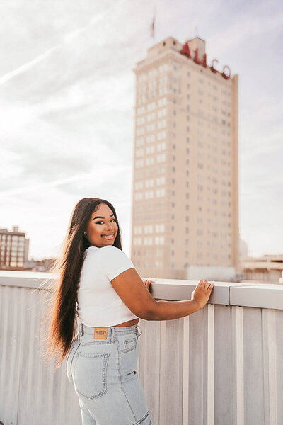 Smiling senior poses on a rooftop in Waco, TX, with the iconic Alico Building in the background captured by a Waco photographer.