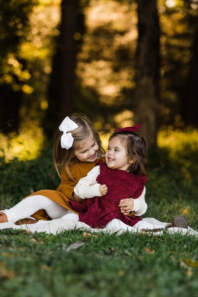 Two young girls playing and hugging each other on a grassy area with autumn foliage in the background, captured by a skilled family photographer in Pittsburgh.