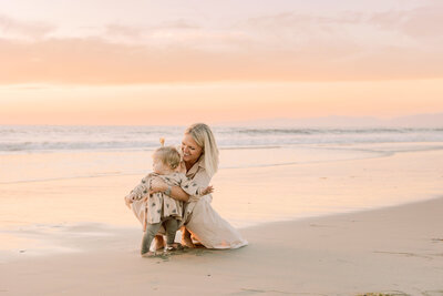 Mom hugging toddler on the beach during a golden morning moment by the ocean.
