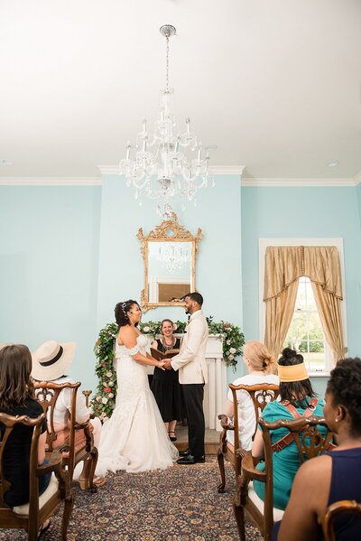 The Afircan American bride and groom recite their vows at their micro wedding in front of a mantle draped with a garland of greenery and flowers